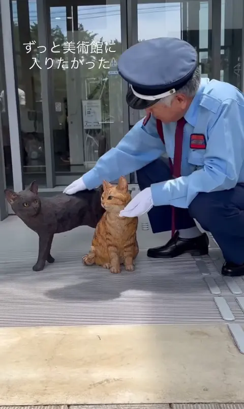 A museum guard crouches at the entrance beside two carved cat figures, a black one honouring Ken-chan and an orange tabby honouring Go-chan, placed just inside the doors.
