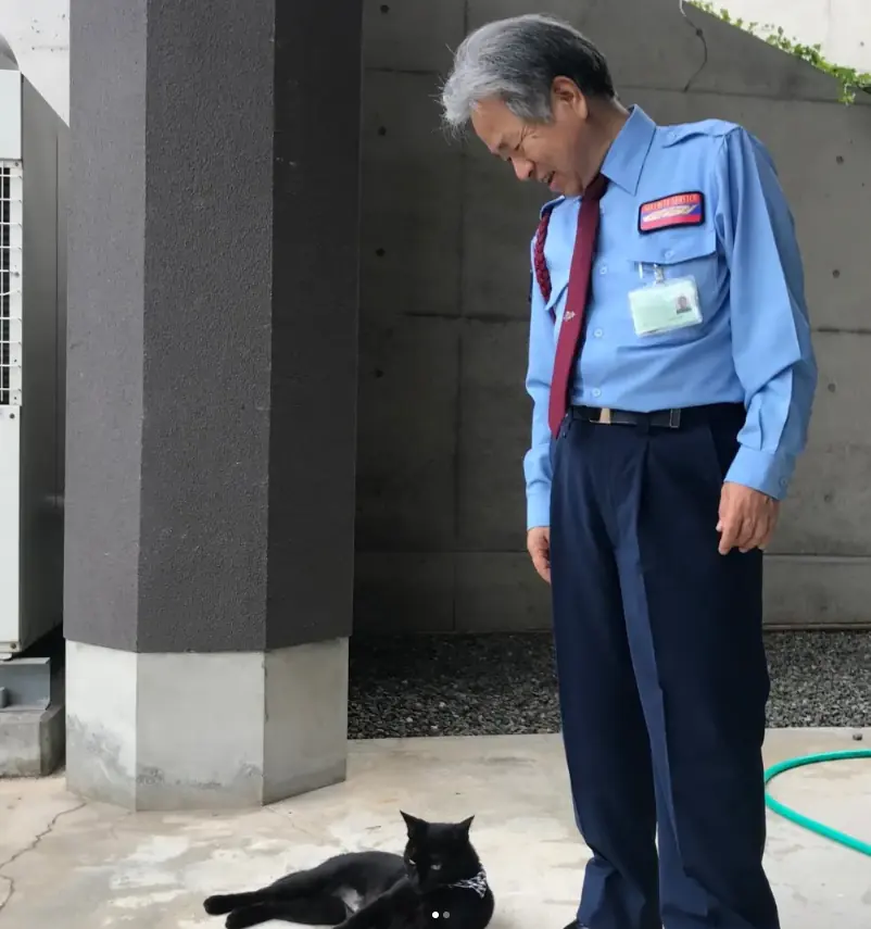 A silver-haired museum guard in a light blue uniform shirt stands on a concrete path, looking down at Ken-chan who sits calmly at his feet.